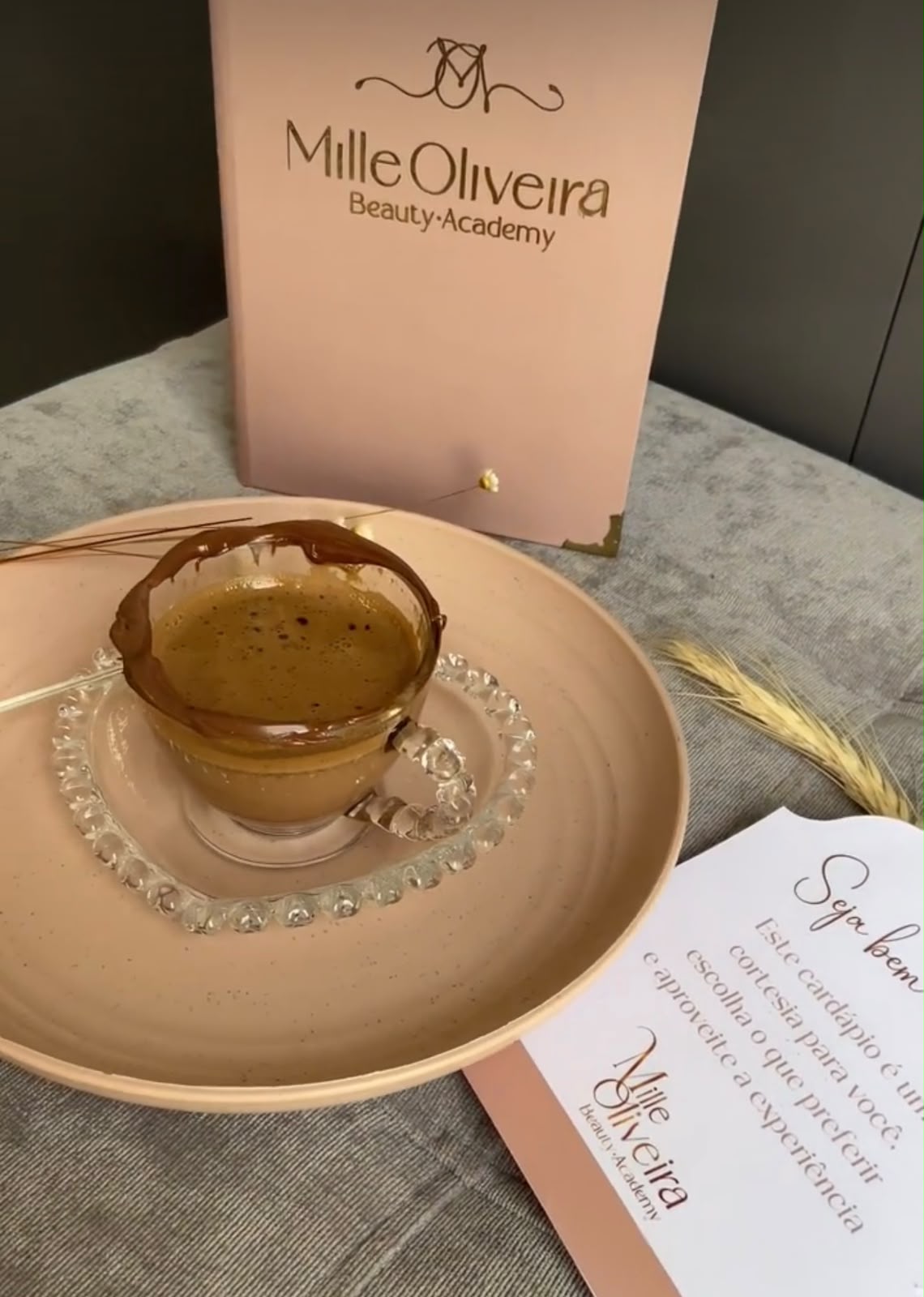 Café servido em xícara de vidro sobre prato rosa, ao lado do cardápio de cortesia da Mille Oliveira